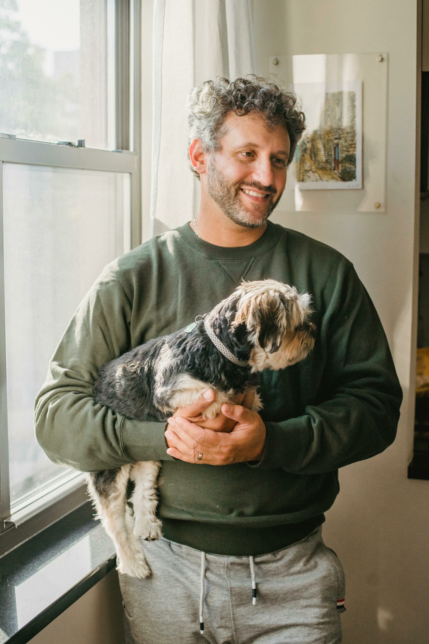 Adult man with ennui holding purebred dog while standing near window